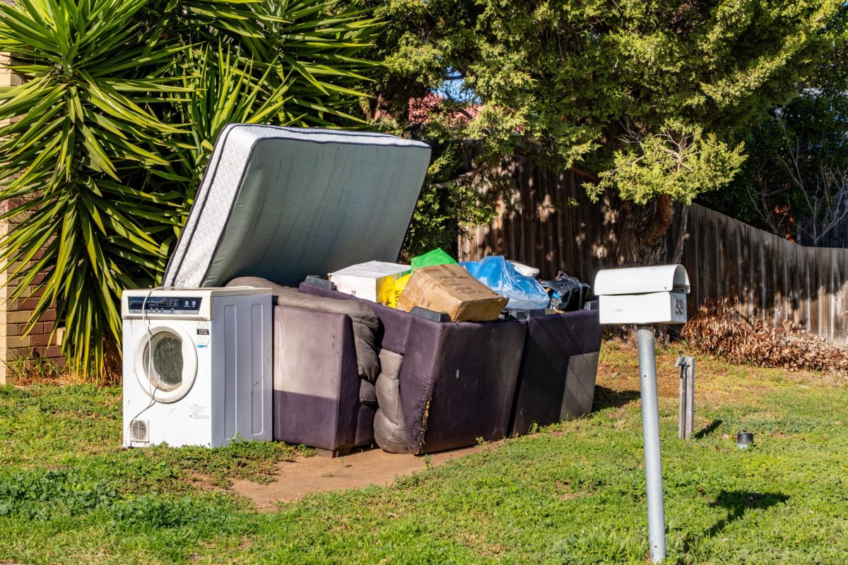 Hard rubbish left on the front yard garden of a suburban property featuring an old washing machine, broken furniture, mattresses, and discarded household items. Urban waste disposal in Australia.